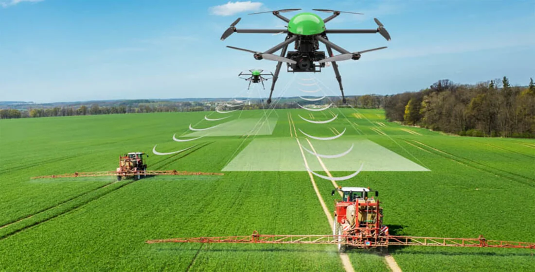 A drone surveying a large field with tractors on the ground spraying crops.